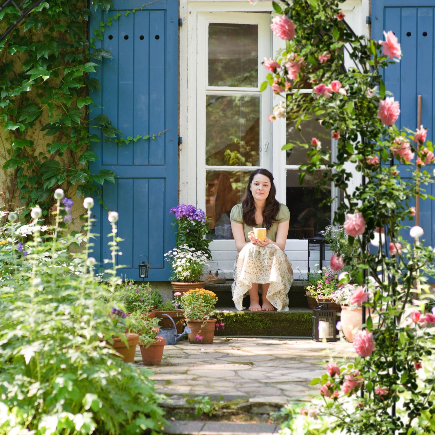 Romantischer Garten mit Rosenbogen, bewachsen mit Rosen, und einer Frau vor blauem Fenster.