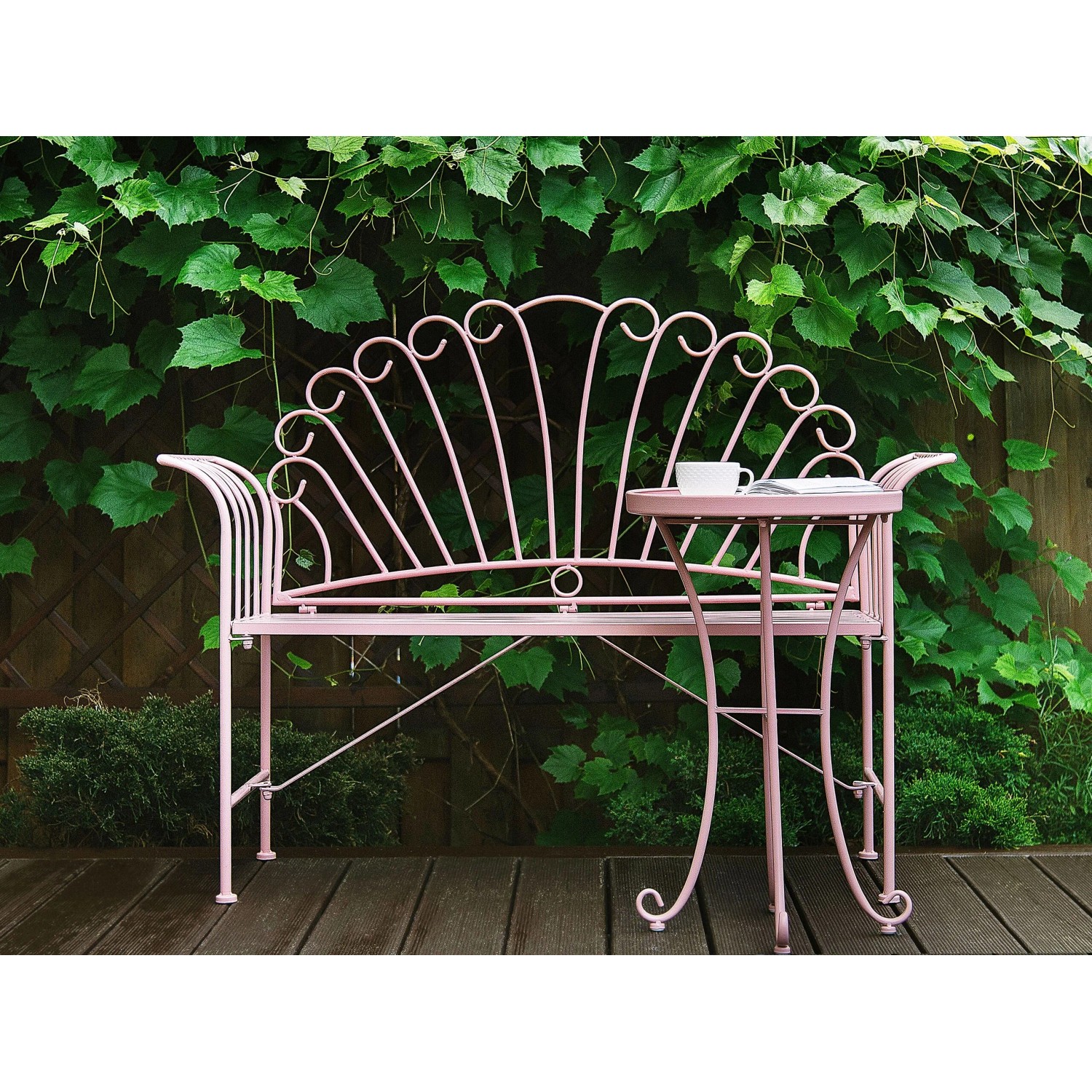 Rosafarbenes Metall Bistro Set CAVINIA mit verzierter Gartenbank und Beistelltisch auf Holzterrasse vor grünen Pflanzen.