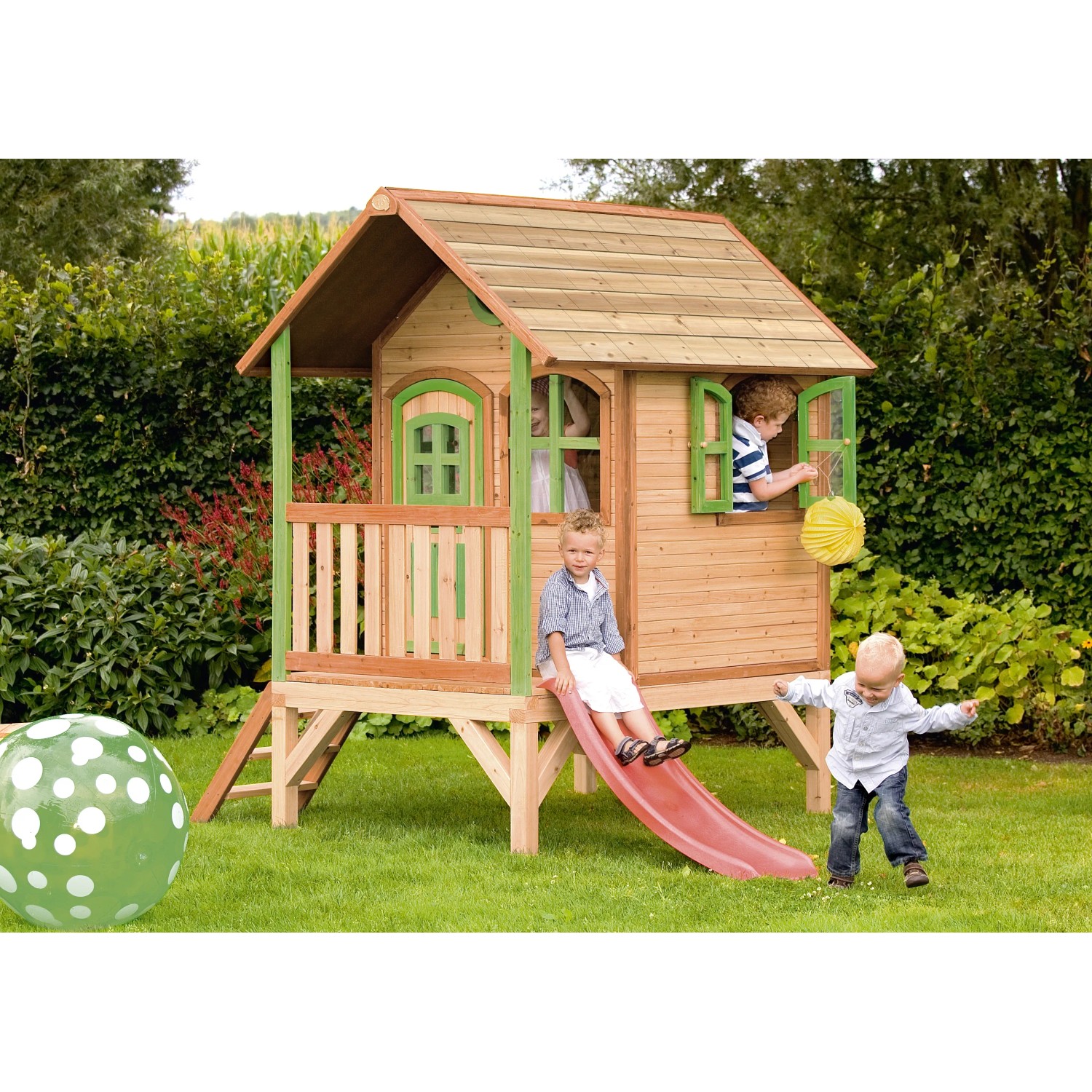Axi Tom Spielhaus aus Holz mit roter Rutsche und spielenden Kindern im Garten.