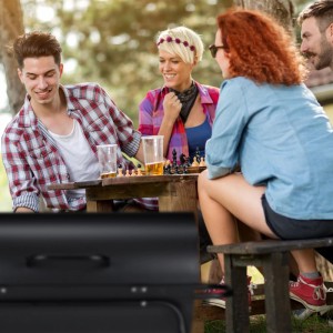 Schwarzer Relaxdays Holzkohlegrill mit Deckel und Rollen im Vordergrund; Freunde spielen im Garten Schach.