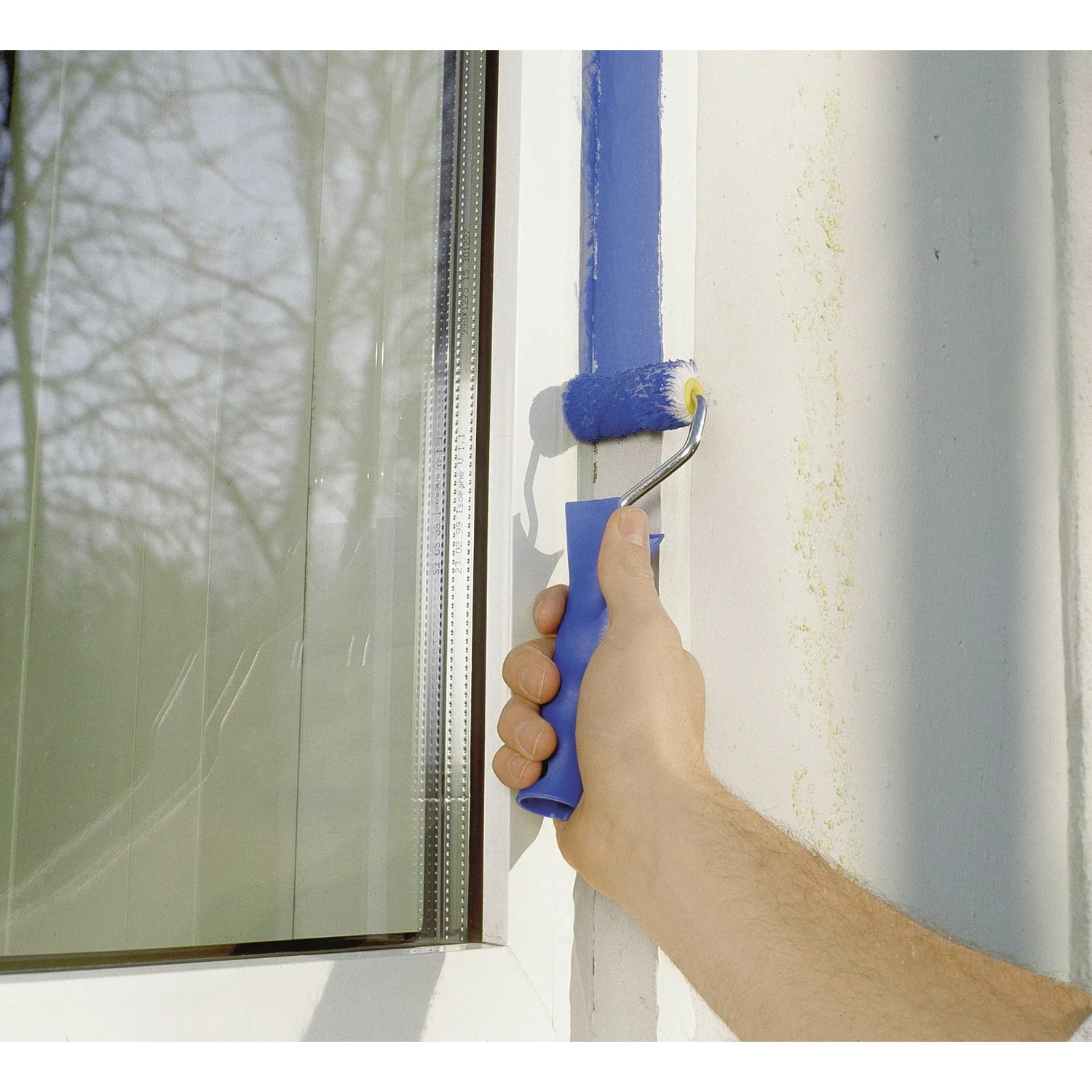 Hand streicht mit LUX Goldfadenwalze (5 cm) blaue Farbe an eine Wand neben einem Fenster.