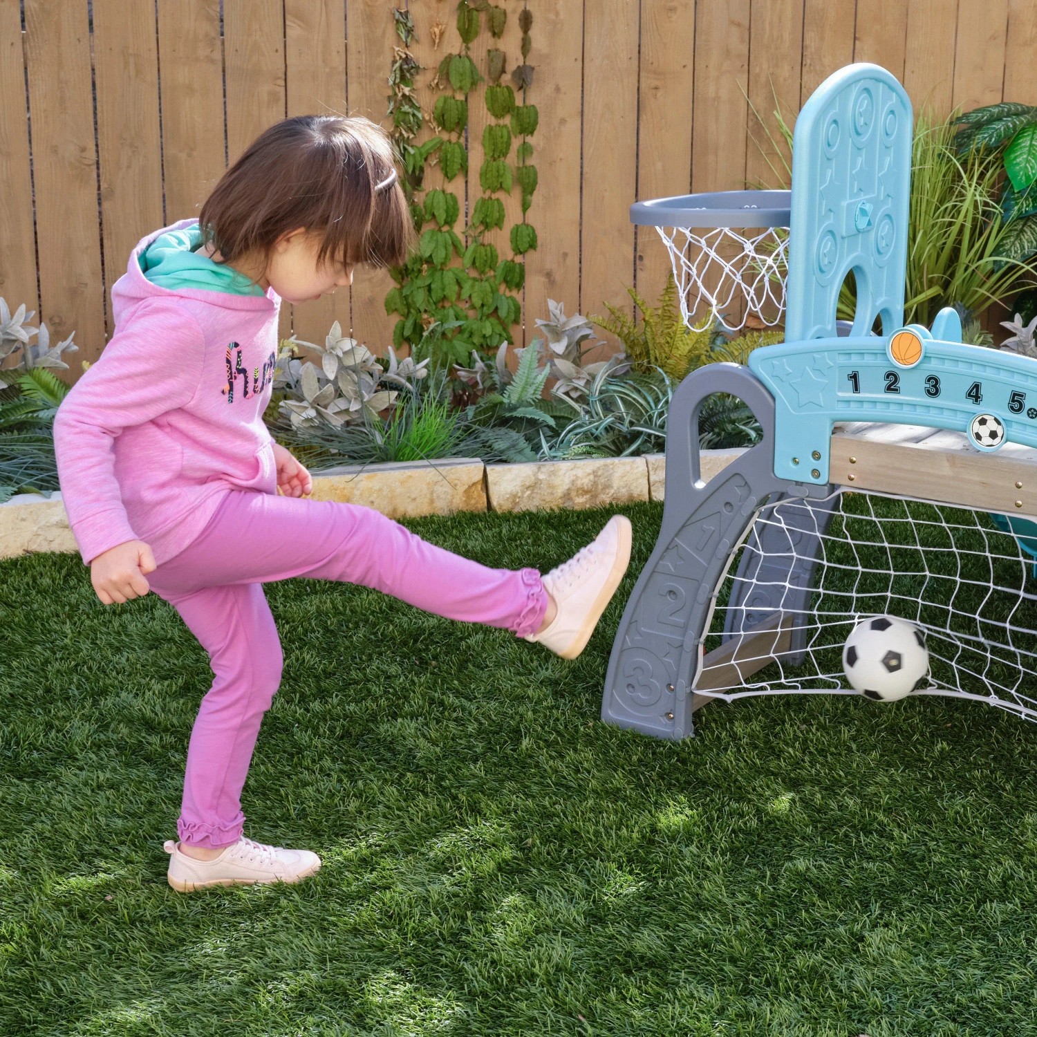 Kind spielt Fußball am KidKraft 5 in 1 Klettergerüst mit Rutsche, Fußballtor & Basketballkorb im Garten.