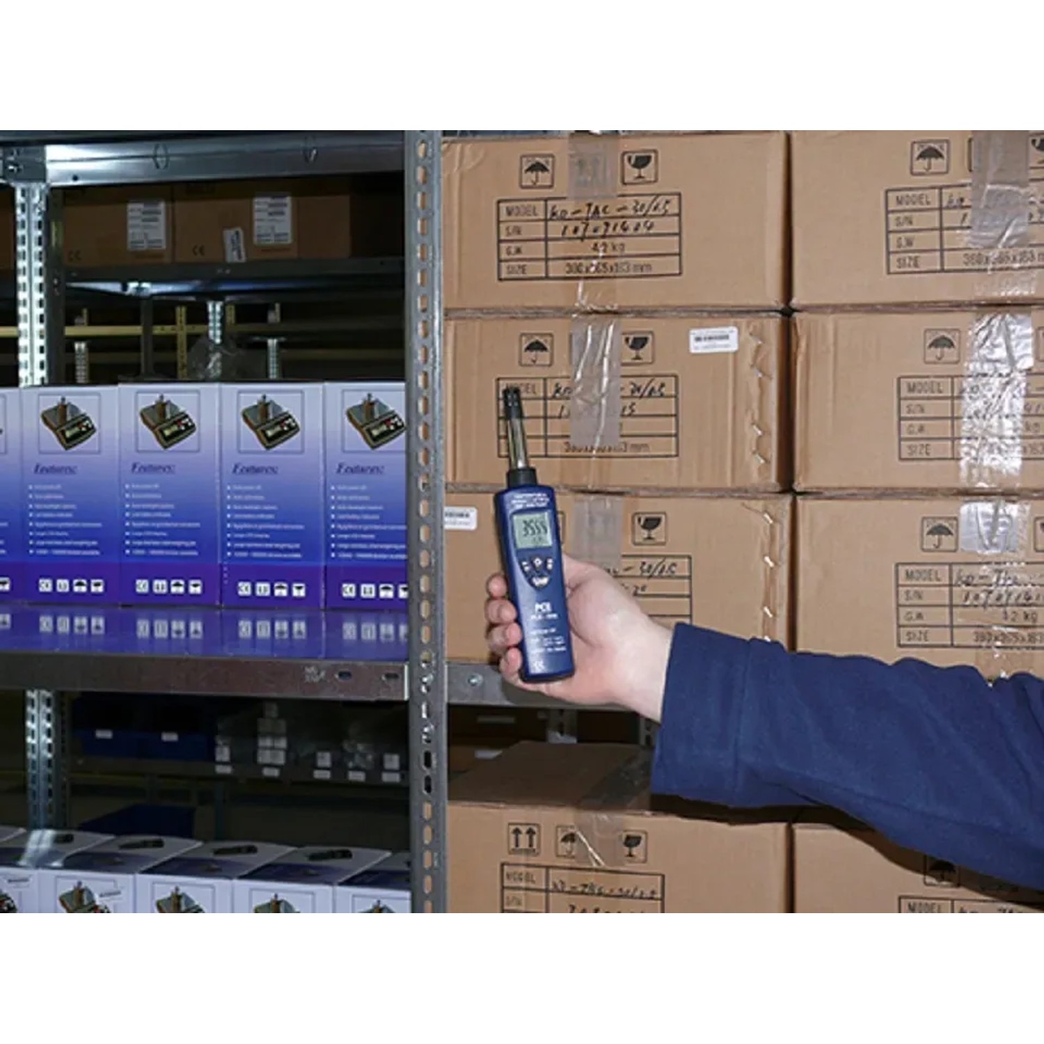 PCE Instruments Hygrometer 555 in Hand vor Lagerregal mit Kartons. Messwerkzeug zur Feuchtigkeitsmessung.