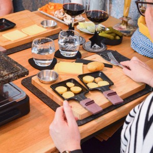 Raclette Teller Set aus Bambus mit Dipschale für 6 Personen. Holzbrett mit Pfännchen, Käse und Beilagen am Tisch.