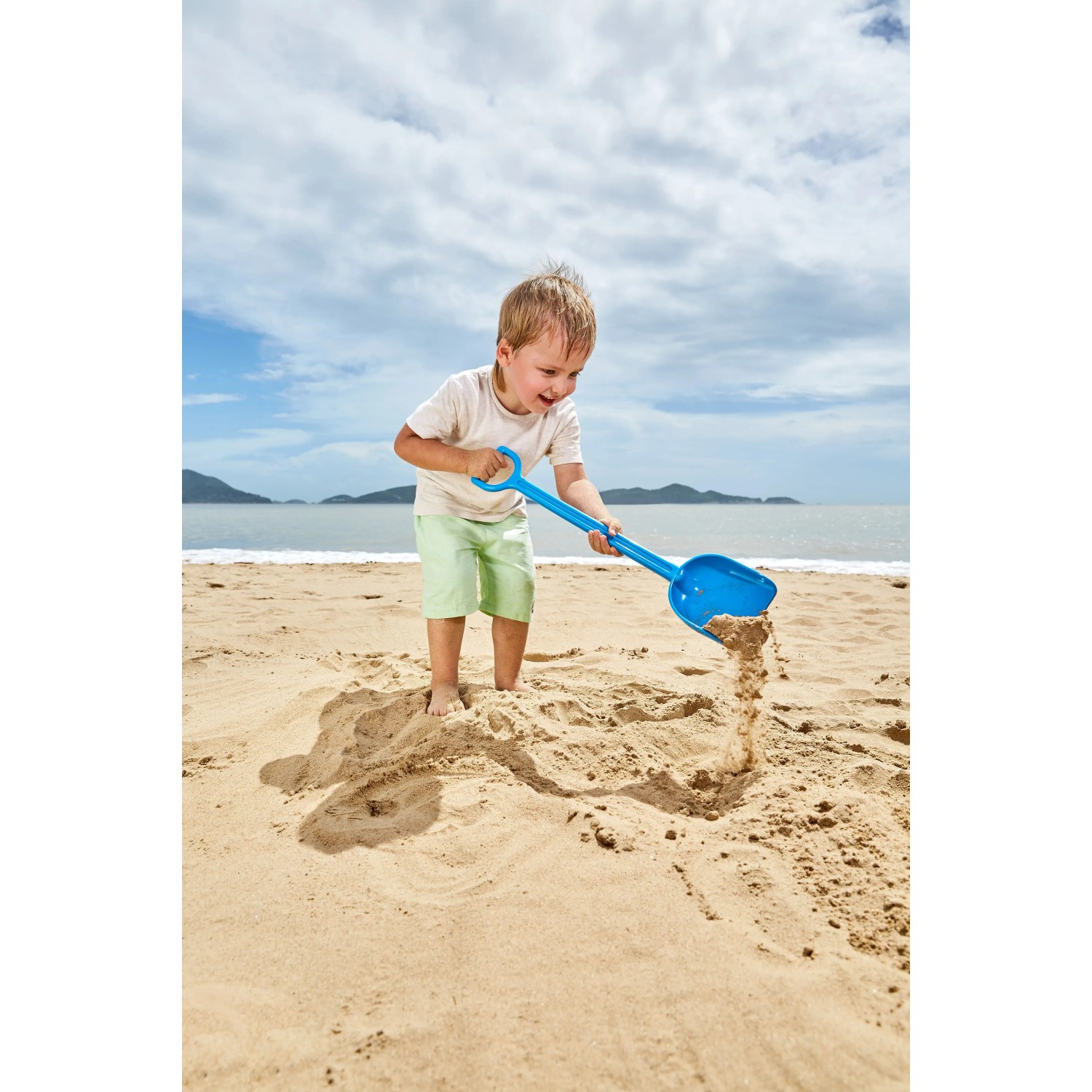 Kind schaufelt Sand mit blauer Hape Sandschaufel am Strand. Sandspielzeug für Spiel & Spaß.