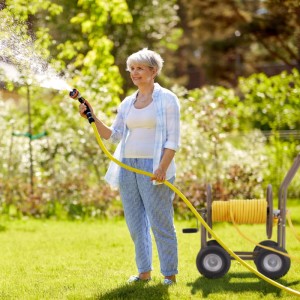 Frau bewässert Garten mit gelbem Schlauch und mobilem Relaxdays Schlauchwagen Stahl Braun mit 4 Rädern.