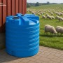 Großer blauer KMND 8000L Trinkwasser Wassertank aus PE mit Rippen, auf Pflaster neben rotem Gebäude, Schafweide.