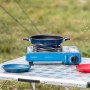 Campingaz Camp Bistro DLX Tischkocher mit Pfanne, Teller und Blumenvase auf einem Campingtisch.