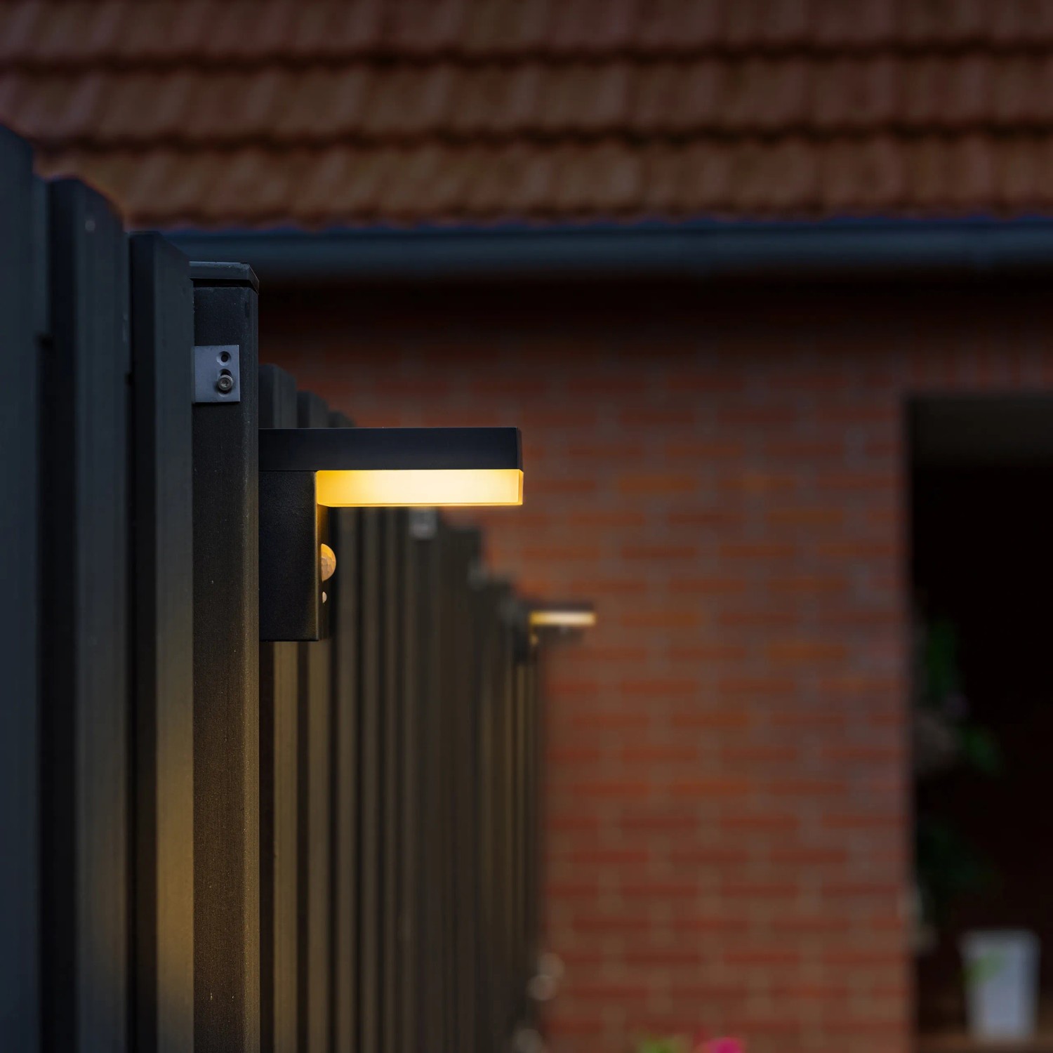 Schwarze Solar-Wandleuchte mit Sensor an einem Zaun. Warmweißes Licht.