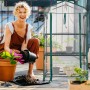 Frau pflanzt auf Balkon. Grünes Balkongewächshaus mit transparentem PVC-Bezug, aufgerollter Tür, 3 Etagen und Anzuchtpflanzen.