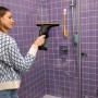 Frau nutzt Kärcher Akku-Fensterreiniger WV 1 Black Edition in Dusche mit lila Fliesen.