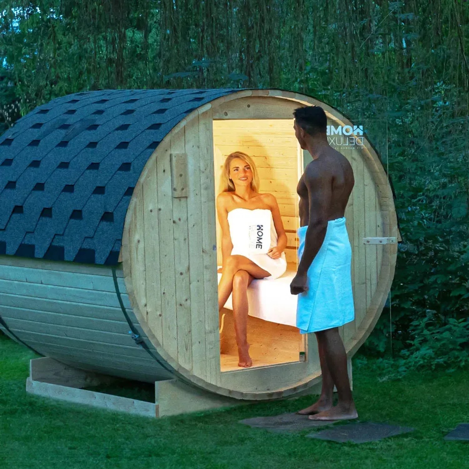 Home Deluxe Fasssauna Lahti Deluxe XL mit Paar vor Holztür im Garten.