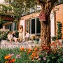 Elho Vogelhaus Cosy, rot-weiß, an Baum befestigt. Gartenszene mit Blumen und entspannten Menschen.