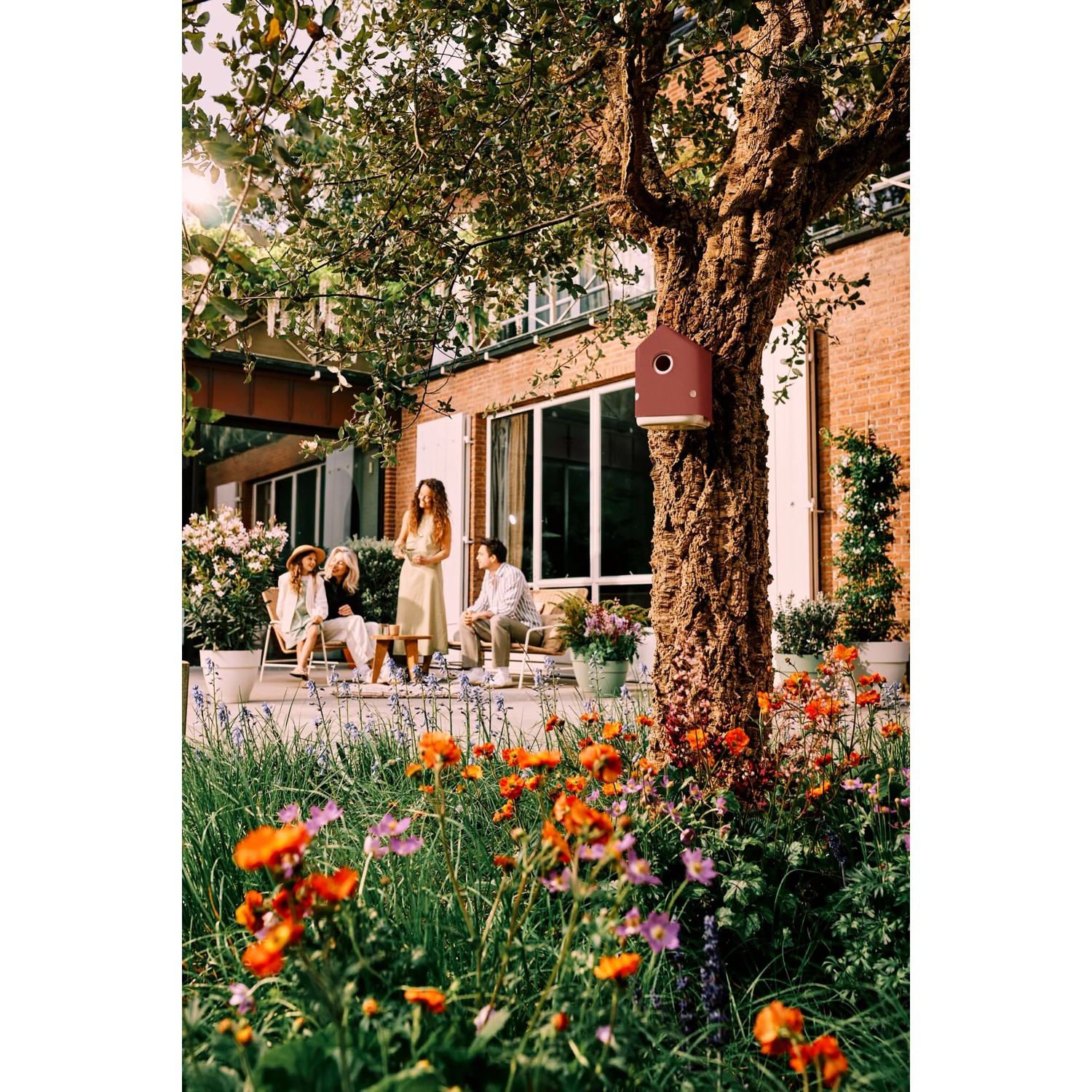 Elho Vogelhaus Cosy, rot-weiß, an Baum befestigt. Gartenszene mit Blumen und entspannten Menschen.
