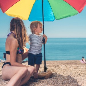 Mutter und Kind am Strand unter buntem Sonnenschirm. Die Relaxdays Bodenhülse sichert den Schirmständer fest im Sand.