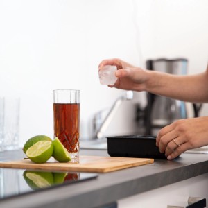 Hand legt großen Eiswürfel aus Silikonform in Glas mit Getränk. Limetten, Küche. Perfekt für XXL Eiswürfel.