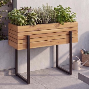 Modernes Hochbeet aus hellbraunem Akazienholz mit dunklen Metallfüßen, bepflanzt mit Kräutern auf der Terrasse.