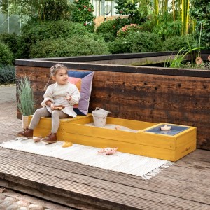 Ockerfarbener MUDDY BUDDY Sandkasten Space Saver mit Kind und Spielzeug auf Terrasse.