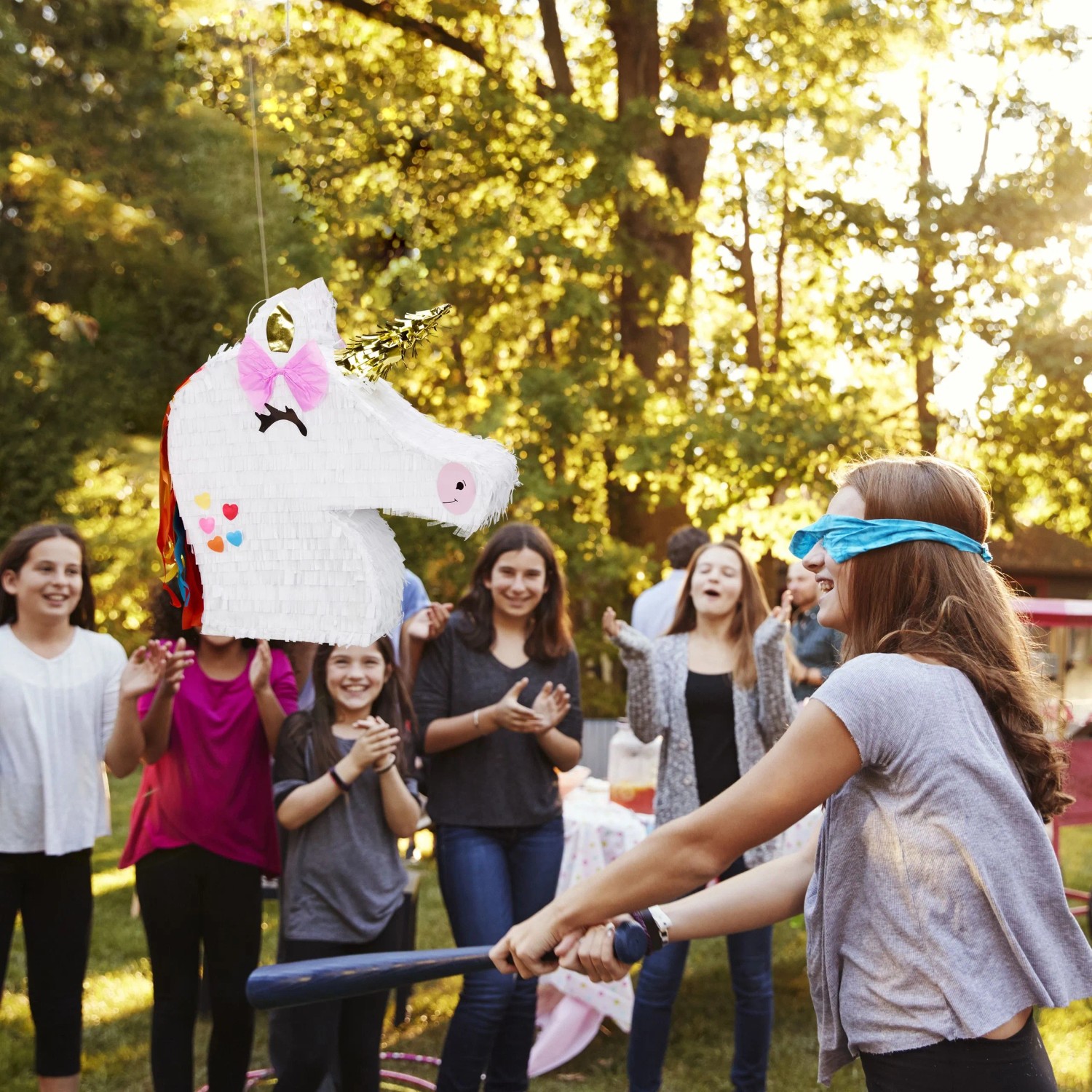 Weiße Einhorn Pinata auf Kindergeburtstag. Blindes Mädchen schlägt mit Schläger auf die Pinata, andere Kinder jubeln. Party Spiel.