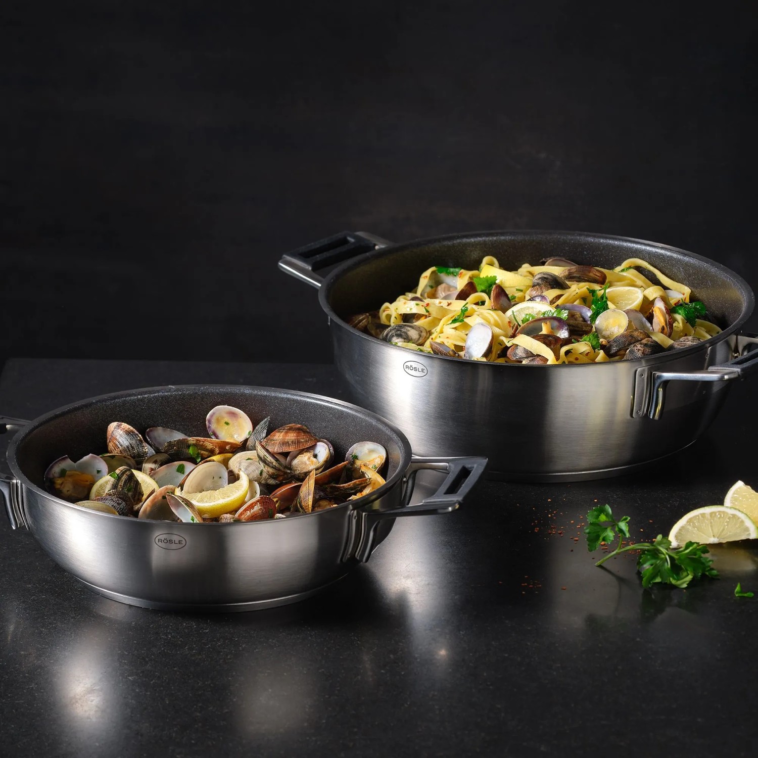 Zwei RÖSLE Servierpfannen SILENCE PRO aus Edelstahl mit ProResist Antihaftbeschichtung, gefüllt mit Pasta und Muscheln, bereit zum Servieren.