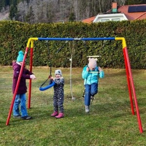 Kettler Doppelschaukel Combi Rot/Blau aus Metall mit Schaukelsitz und Leiter. Kinder spielen im Garten.
