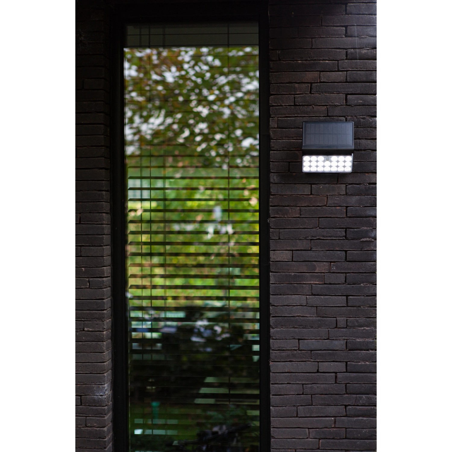 Schwarze Lutec LED Solar-Wandleuchte Tuda mit Bewegungsmelder an dunkler Backsteinwand.