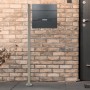 ML-Design Standbriefkasten mit Zeitungsfach und silbernem Edelstahl-Pfosten vor einer Backsteinwand.