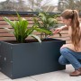 Herstera Garden Pflanzkasten, anthrazit, 150x50x50 cm mit Bepflanzung und Frau bei der Gartenarbeit.