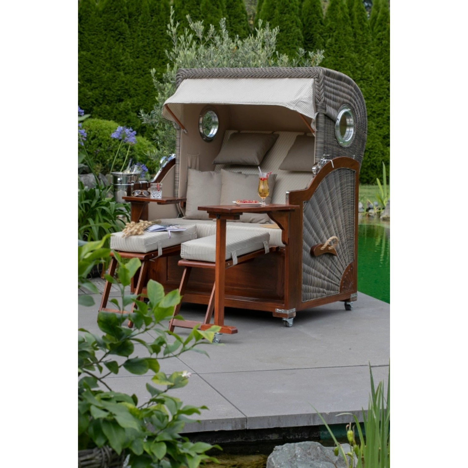 Strandkorb Westerland Mahagoni mit Bullauge, grauem PE-Geflecht und Markise im Garten.