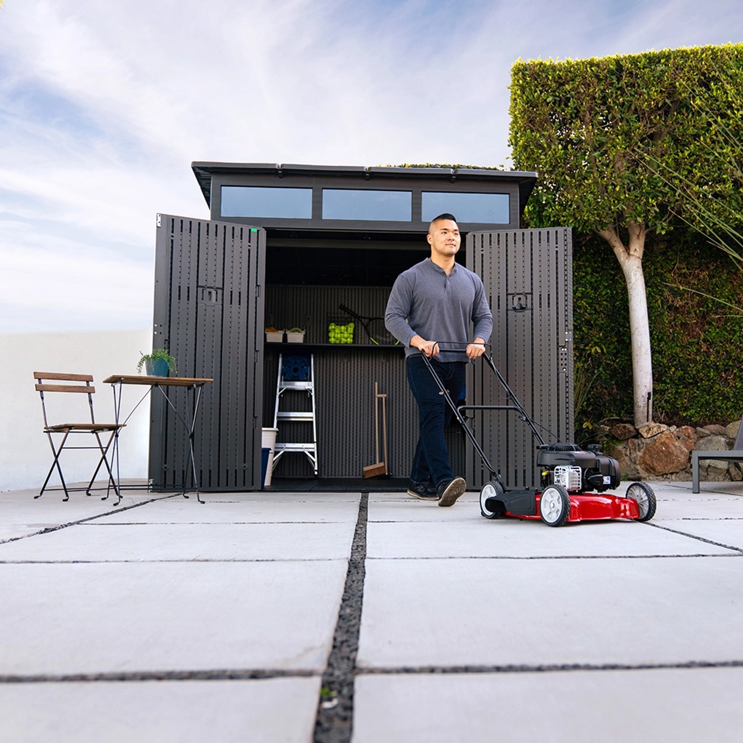 Lifetime Gerätehaus Merkur dunkelgrau (5,4 m²) mit Mann und Rasenmäher vor geöffneter Tür.