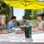 Zwei Bosch UniversalSpeaker 18V-1 Bluetooth-Lautsprecher auf Holztisch, Menschen genießen Musik im Garten.