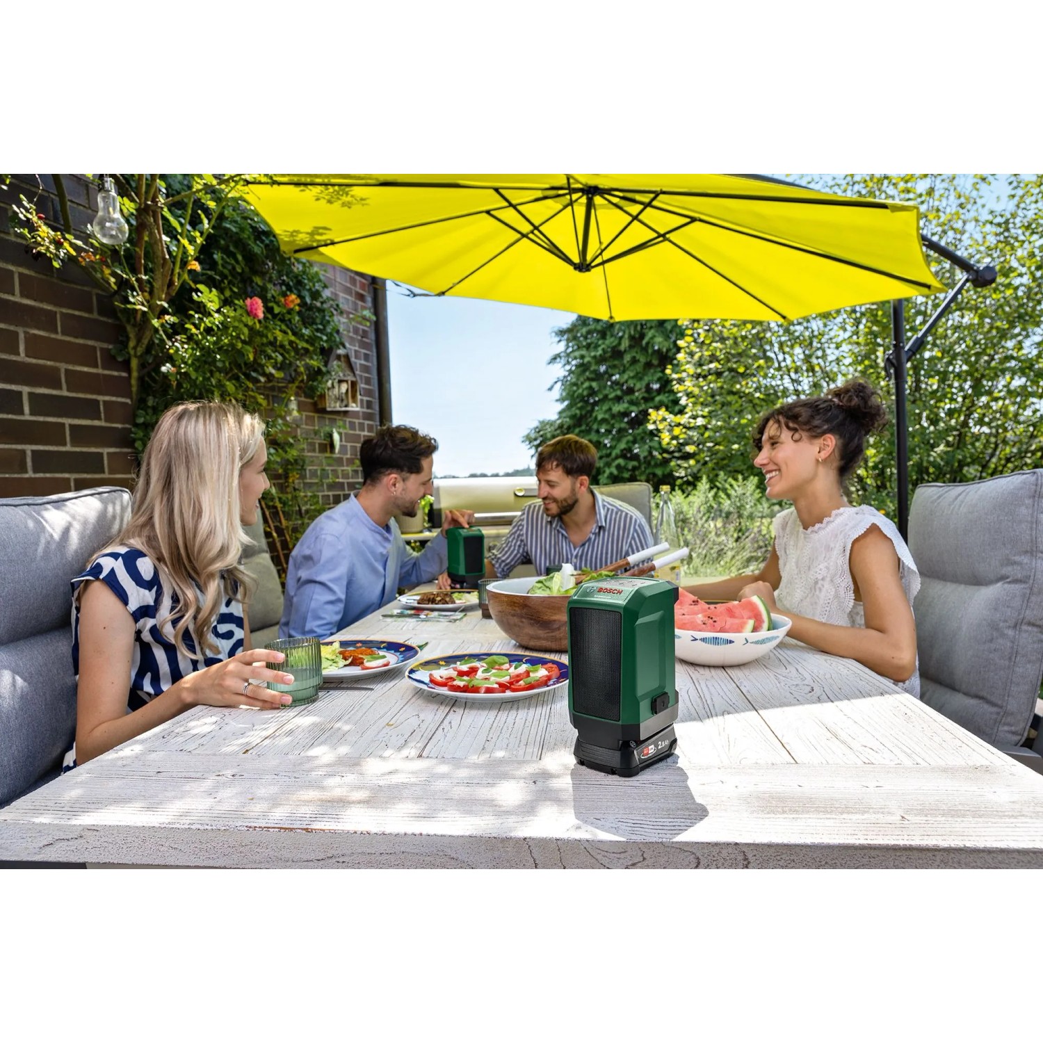 Zwei Bosch UniversalSpeaker 18V-1 Bluetooth-Lautsprecher auf Holztisch, Menschen genießen Musik im Garten.