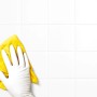 Hand mit weißem, latexfreiem Einmalhandschuh reinigt weiße Fliesen mit gelbem Tuch.