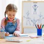 Lachendes Kind malt am Tisch. Im Hintergrund steht eine höhenverstellbare Relaxdays Holz Standtafel mit Whiteboard für Kinder.