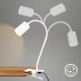 Weiße Briloner LED-Klemmleuchte FLEKS mit vier schwenkbaren Leuchtenarmen am Tisch befestigt.