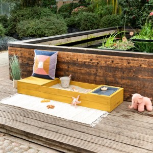 Ockerfarbener MUDDY BUDDY Sandkasten Space Saver mit Spielzeug und Wasserwanne auf Terrasse.