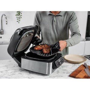 Ninja Foodi Grill AG301EU: Grau-schwarzer Grill mit Steaks auf Grillplatte, bedient von Person mit Grillhandschuhen.