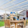 Helle Gutta Terrassenüberdachung Premium, weiße Stegplatten, Gartenmöbel und Spielplatz im Hintergrund.