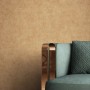 Goldene, strukturierte Vliestapete Zeno mit feiner Textiloptik an Wand, daneben moderner Sessel.