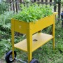 Gelbes Herstera Garden Metall-Hochbeet mit Rädern und Ablagefläche, bepflanzt mit Kräutern.