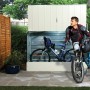 Duramax Fahrradbox in Hellbeige mit zwei Fahrrädern und einer Person davor im Garten.