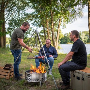 Petromax Dreibein mit Grillrost über Feuerstelle, umgeben von drei Personen in der Natur.