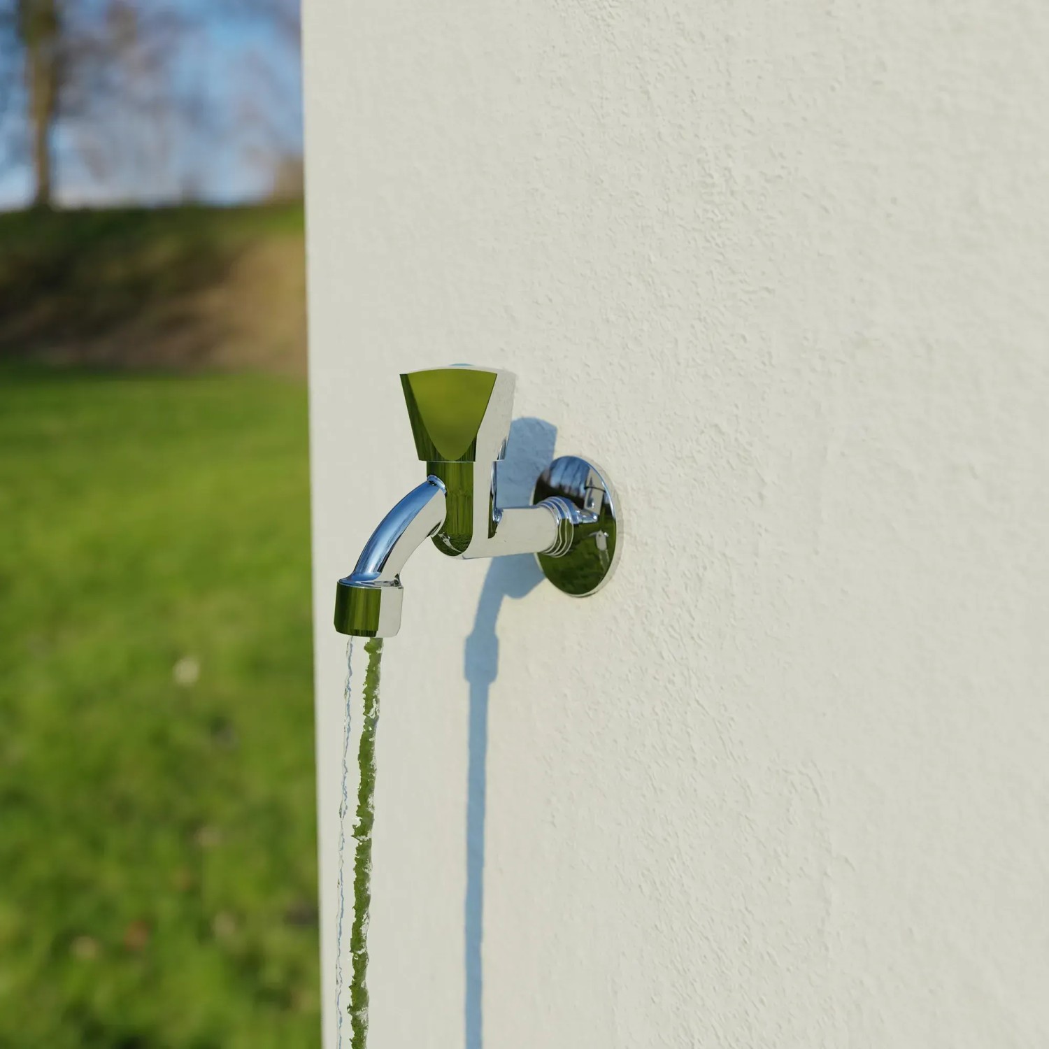 Chromfarbenes Kirchhoff Auslaufventil mit Haubengriff und Wasserstrahl vor weißer Wand.