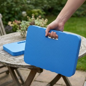 Hand hält blaues Relaxdays Kniekissen aus EVA-Schaumstoff mit Griff. Ein weiteres Kissen liegt auf Gartentisch. Knieschutz 2er Set.