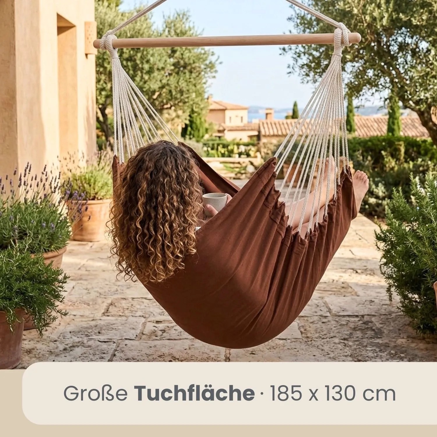 Brauner AMANKA Hängesessel aus Baumwolle. Eine Person entspannt auf einer Terrasse mit Pflanzen. Ideal für Garten & Outdoor.