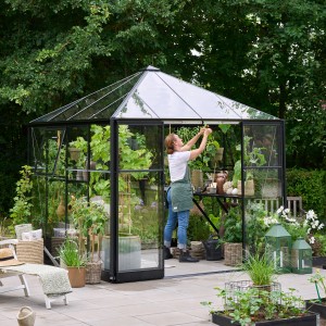 Schwarzes Halls Gewächshaus Qube Square mit Sicherheitsglas, genutzt von einer Person im Garten.